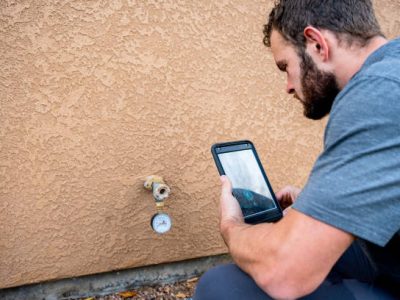 Young Male Property Inspector Testing Water Pressure and Photographing an outside Water Faucet on a modern brown stucco home